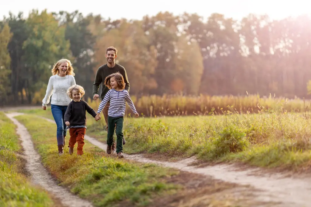 A family of four enjoying a walk in the countryside. The mother and father, dressed in sweaters and jeans, are walking behind their two children, a boy and a girl, who are holding hands and running ahead. The boy is wearing a dark sweater and orange pants, while the girl is wearing a striped shirt and green pants. They are on a dirt path surrounded by green grass and trees with autumn foliage in the background, bathed in warm sunlight.