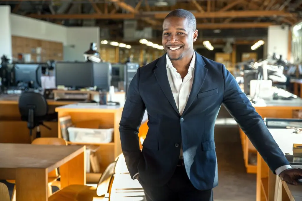 A confident and smiling man in a dark suit jacket and white shirt standing in an office environment with desks and computers in the background.