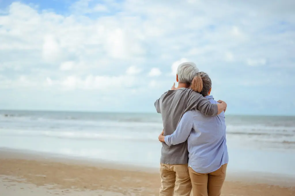 An elderly couple standing on a beach, embracing as they look out over the ocean under a partly cloudy sky.