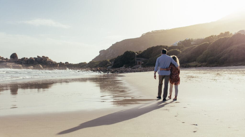 A couple walking arm in arm along a serene beach at sunset. The man is wearing a blue shirt and khaki pants, and the woman is wearing a patterned dress. They are walking close to the water's edge, with the waves gently lapping at the shore. In the background, there are rocky formations and hills covered in greenery, illuminated by the soft, warm light of the setting sun.