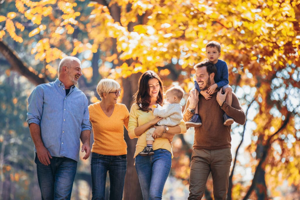 A multi-generational family enjoying a walk in a park during autumn. The grandfather, wearing a blue shirt, is walking beside the grandmother, who is in a yellow sweater. The mother, holding a baby, is also wearing a yellow top and is smiling at the child. The father, in a brown sweater, is carrying another child on his shoulders. They are all walking together under trees with vibrant autumn leaves, enjoying a warm and cheerful moment.