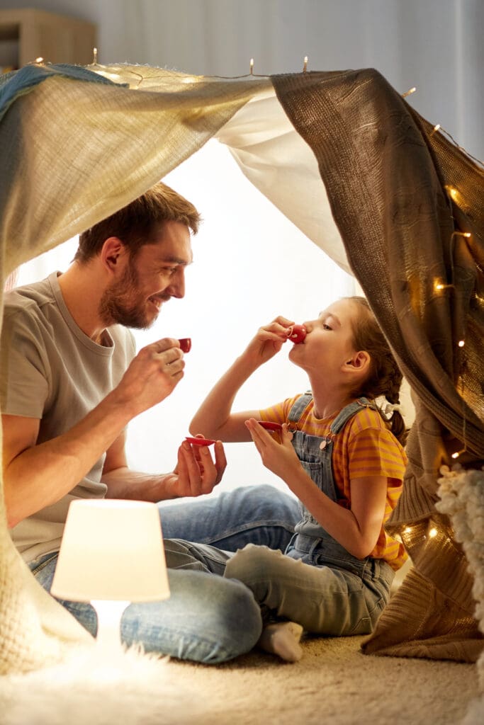 A father and daughter enjoying a pretend tea party inside a cozy blanket fort. The father, wearing a gray t-shirt and jeans, smiles while holding a tiny red teacup. The daughter, wearing a yellow striped shirt and overalls, drinks from a matching teacup. They are sitting on a soft carpet with a small lamp beside them, and the fort is decorated with string lights.