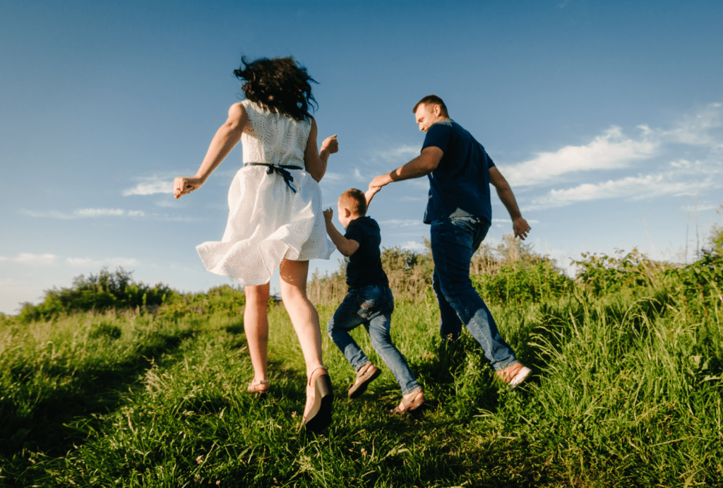 A family of three running together in a grassy field. The mother, wearing a white dress, the father, in a blue shirt and jeans, and their young son, also in a blue shirt and jeans, are holding hands and joyfully running up a hill. The sky is clear with a few clouds, and the scene is filled with the energy and excitement of their activity.