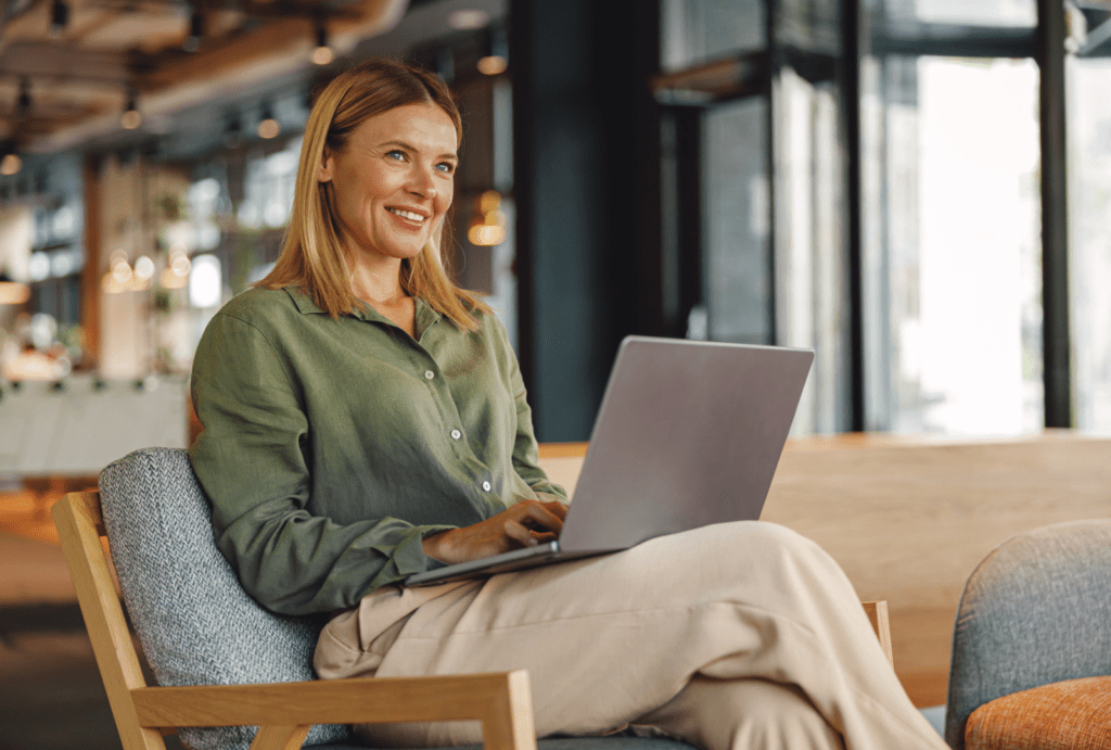 A woman sitting in a modern, well-lit office or cafe, using a laptop. She is smiling and appears engaged with her work. She is wearing a green blouse and beige pants, and is seated on a comfortable chair. The background shows a contemporary interior with large windows, stylish furniture, and soft lighting.