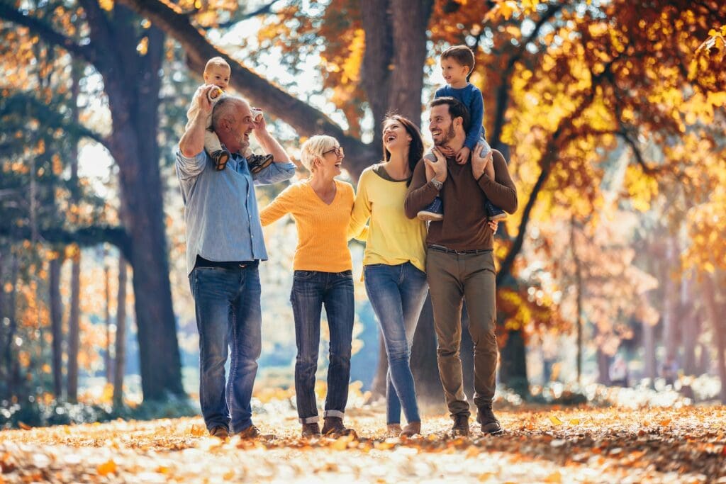 A multi-generational family enjoying a walk in a park during autumn. The grandfather is carrying a baby on his shoulders, the grandmother is walking beside him, smiling. Next to them, a young woman and a man, possibly the parents, are also walking and laughing, with the man carrying a young boy on his shoulders. The scene is filled with autumn leaves and tall trees, creating a warm and cheerful atmosphere.