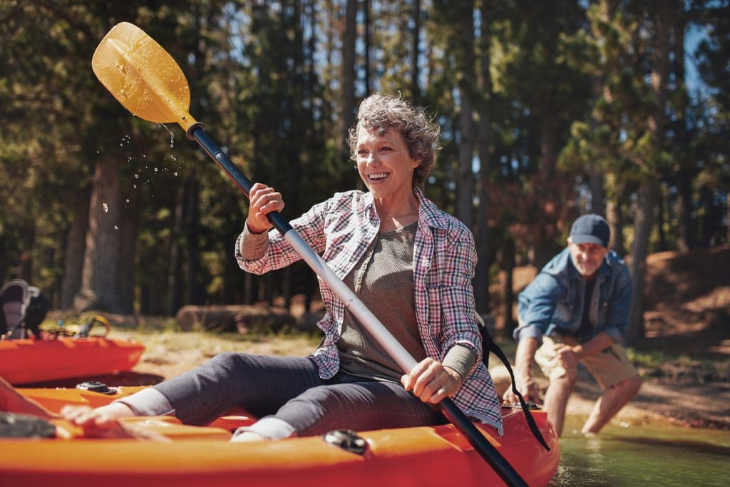 An older woman joyfully kayaking in a bright orange kayak, lifting a yellow paddle that drips with water. She is smiling broadly, dressed in a plaid shirt and gray pants. A man, likely her companion, is visible in the background, also in a kayak. They are surrounded by a serene, forest-lined lake.