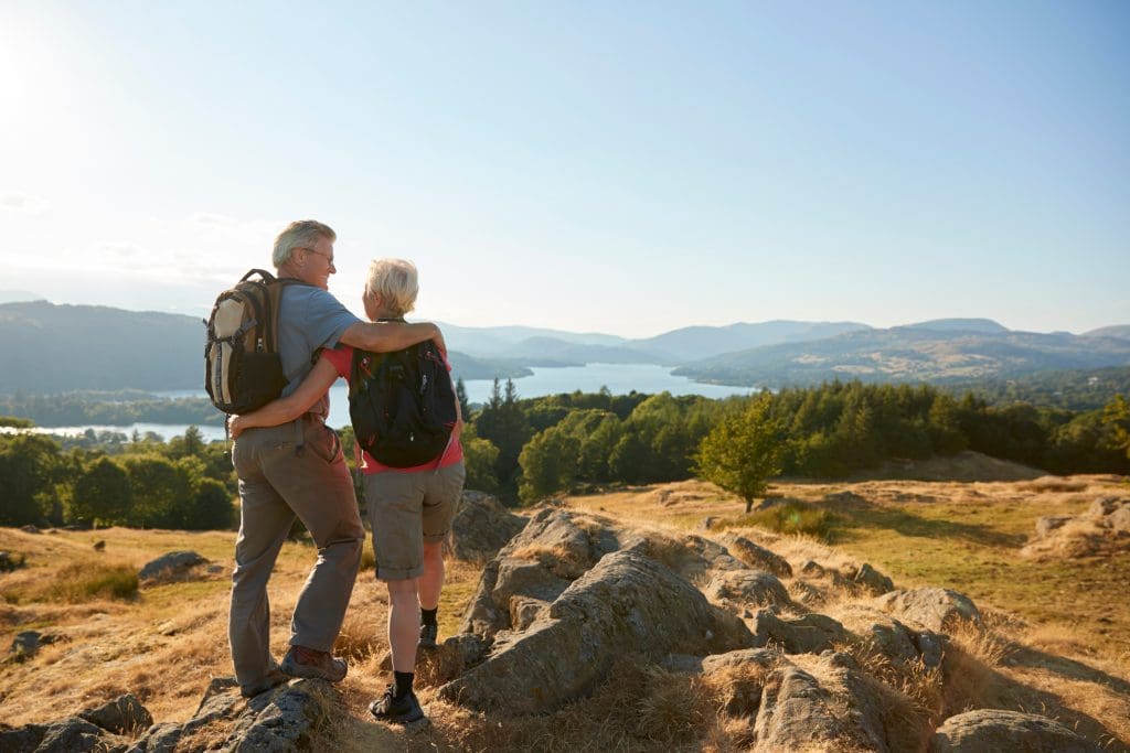 An older couple enjoying a scenic view from a hilltop overlooking a lake and distant mountains during a sunny day. They are embracing while looking out at the landscape, wearing backpacks and hiking attire.